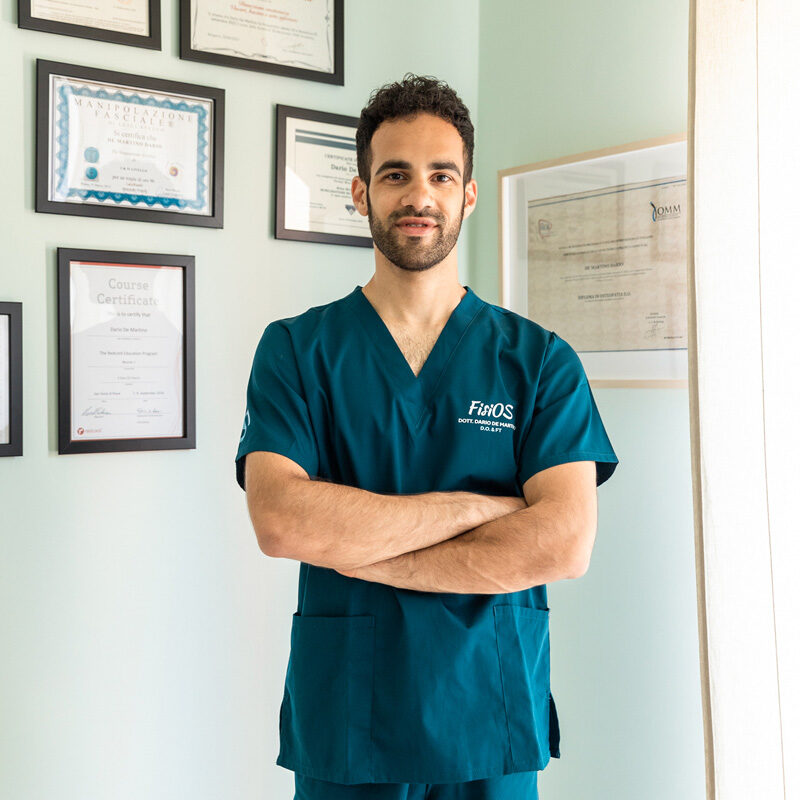 Dottore Dario De Martino osteopata e fisioterapista in piedi nel suo studio con divisa medicale davanti alla parete con le sue certificazioni